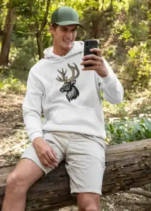 Homme portant un hoodie blanc avec un design de cerf majestueux, assis dans un cadre naturel.