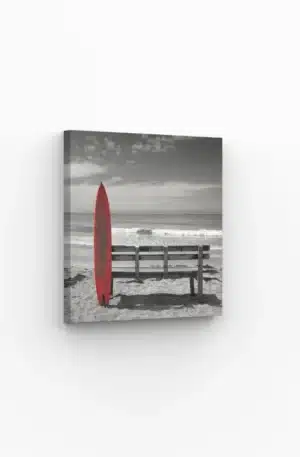 Planche de surf rouge appuyée contre un banc en bois sur une plage déserte.